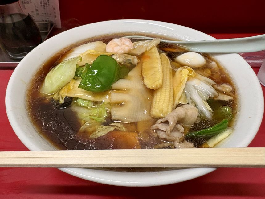 白い楕円皿に盛られた具だくさんのラーメン、エビ・チンゲン菜・ヤングコーンが浮かぶ醤油スープ